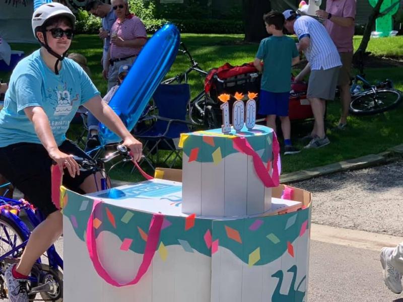 A woman riding the book bike in a parade. The bike is decorated as a centennial birthday cake.